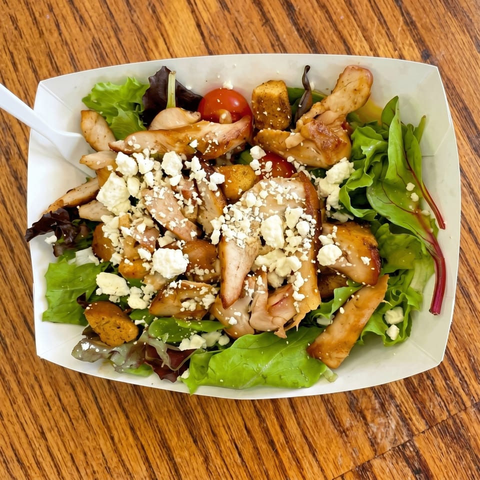 A chicken salad with feta cheese, cherry tomatoes, and croutons in a white paper container.
